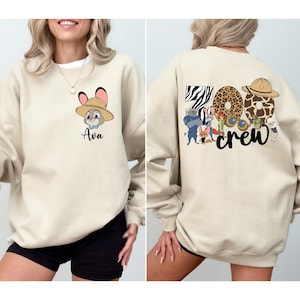 Puede incluir: Sudadera beige con un personaje de dibujos animados con sombrero y el nombre "Ava" debajo. La parte trasera de la sudadera tiene las palabras "Zoo Crew" con dise&ntilde;os de estampado animal y personajes de dibujos animados.