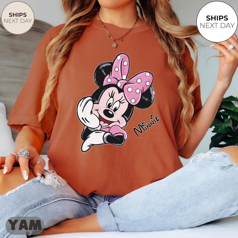 May include: Rust-colored t-shirt featuring a Minnie Mouse graphic. The cartoon character is depicted with a pink bow with white polka dots, a pink dress, and the word "Minnie" in cursive. The shirt is a relaxed fit.