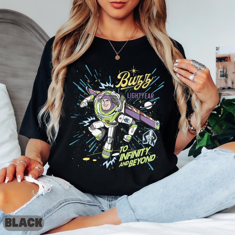 May include: Black t-shirt featuring a graphic of Buzz Lightyear in his spacesuit, with the text "Buzz Lightyear" and "To Infinity and Beyond." The design includes a space background with stars and planets. The word "BLACK" is printed at the bottom.