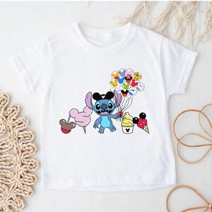 Puede incluir: Camiseta blanca con un personaje de dibujos animados de Stitch con un sombrero de Mickey Mouse y globos. El dise&ntilde;o incluye algod&oacute;n de az&uacute;car, helado y otros elementos tem&aacute;ticos de Disney. Un dise&ntilde;o divertido y colorido.