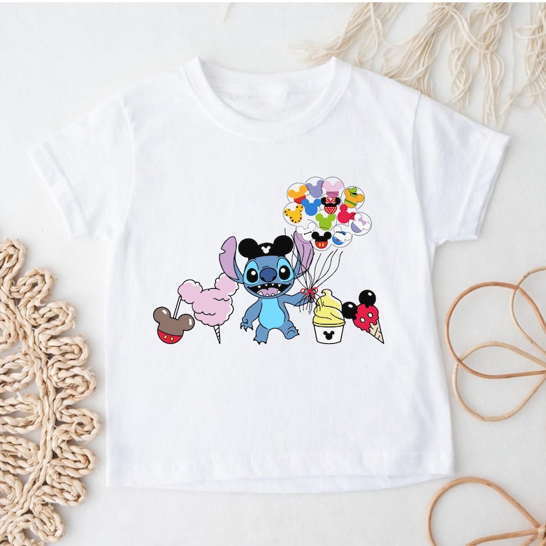 Puede incluir: Camiseta blanca con un personaje de dibujos animados de Stitch con un sombrero de Mickey Mouse y globos. El dise&ntilde;o incluye algod&oacute;n de az&uacute;car, helado y otros elementos tem&aacute;ticos de Disney. Un dise&ntilde;o divertido y colorido.