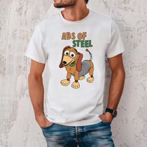 K&ouml;nnte beinhalten: Wei&szlig;es T-Shirt mit einer Cartoon-Illustration von Slinky Dog und dem Text "ABS OF STEEL". Das Design umfasst braune, beige und graue Farben. Das T-Shirt hat einen Rundhalsausschnitt.