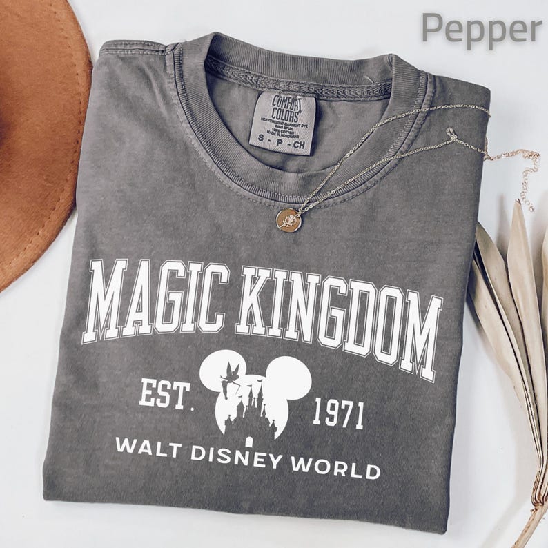 May include: A gray t-shirt with the words "MAGIC KINGDOM" in white block letters. Below the text is "EST. 1971" and "WALT DISNEY WORLD" with a Mickey Mouse silhouette. A gold necklace rests on the shirt.