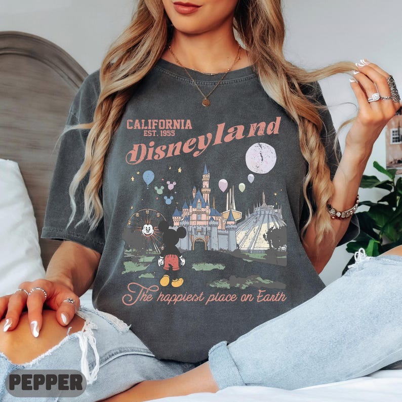 May include: Dark gray t-shirt featuring a Disneyland graphic. The design includes the text "California Disneyland Est. 1955" and an illustration of the castle, Mickey Mouse, and other park elements. The phrase "The happiest place on Earth" is also included.