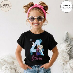 May include: Black t-shirt with a blue number 4, featuring cartoon characters Anna and Elsa from Frozen. The name "Olivia" is written below in cursive. The shirt is designed for a child's birthday celebration.