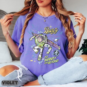 Może przedstawiać: Fioletowa koszulka z grafiką Buzza Astrala w jego skafandrze kosmicznym, z napisem "Buzz Lightyear" i "To Infinity and Beyond". Projekt zawiera planetę i tło o tematyce kosmicznej.