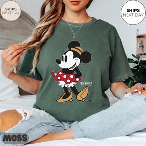 Puede incluir: Camiseta verde con un gr&aacute;fico de Minnie Mouse. El personaje de dibujos animados se representa en una pose cl&aacute;sica, con una falda roja de lunares y zapatos amarillos. La palabra "Disney" est&aacute; impresa debajo.