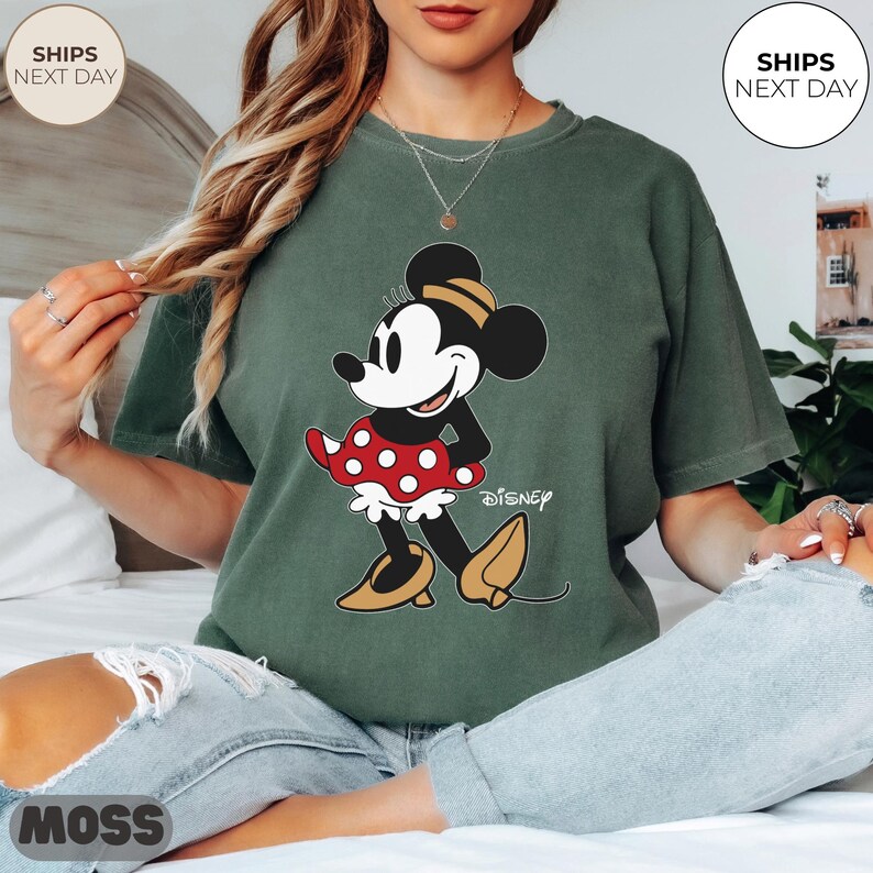 Puede incluir: Camiseta verde con un gr&aacute;fico de Minnie Mouse. El personaje de dibujos animados se representa en una pose cl&aacute;sica, con una falda roja de lunares y zapatos amarillos. La palabra "Disney" est&aacute; impresa debajo.