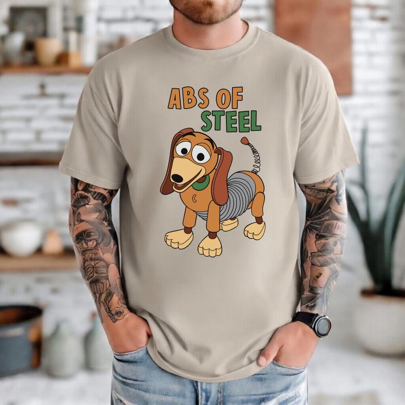 K&ouml;nnte beinhalten: Beiges T-Shirt mit einer Cartoon-Illustration von Slinky Dog und dem Text "ABS OF STEEL" in Orange und Gr&uuml;n. Die Grafik zeigt Slinky Dog mit braunen Ohren, einem gr&uuml;nen Kragen und einem spiralf&ouml;rmigen K&ouml;rper. Dies ist ein Freizeitkleidungsst&uuml;ck.