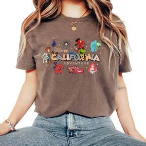 K&ouml;nnte beinhalten: Ein braunes Kurzarm-T-Shirt mit einem Grafikdruck und dem Text Disney CALIFORNIA ADVENTURE. Das Design zeigt verschiedene Zeichentrickfiguren wie Woody, Buzz Lightyear, Baymax, Lightning McQueen, Ariel, Sulley, Mike Glotzkowski und Goofy.
