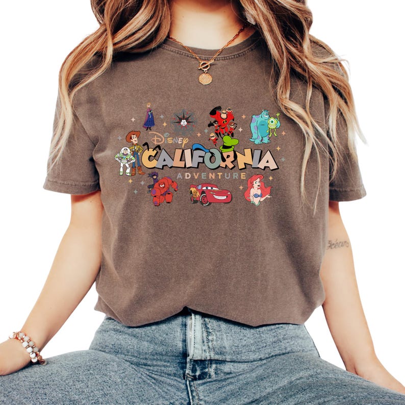 K&ouml;nnte beinhalten: Ein braunes Kurzarm-T-Shirt mit einem Grafikdruck und dem Text Disney CALIFORNIA ADVENTURE. Das Design zeigt verschiedene Zeichentrickfiguren wie Woody, Buzz Lightyear, Baymax, Lightning McQueen, Ariel, Sulley, Mike Glotzkowski und Goofy.