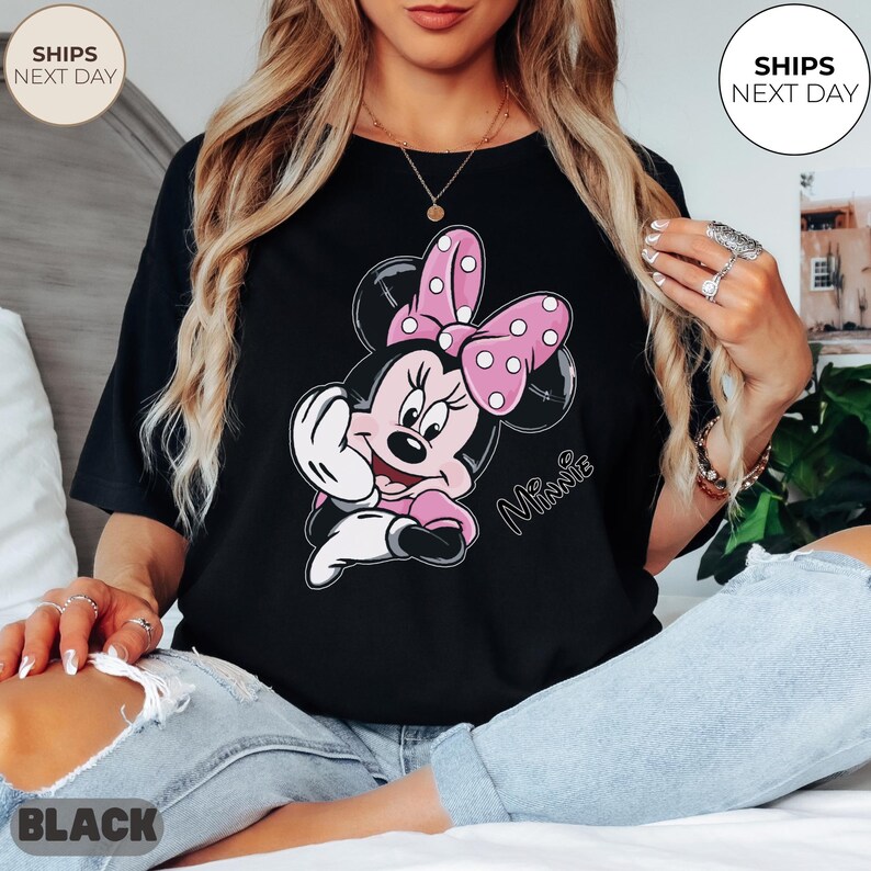 May include: Black t-shirt featuring a cartoon Minnie Mouse design. Minnie Mouse is depicted with a pink bow with white polka dots, a pink dress, and a white glove. The word "Minnie" is written in cursive. The shirt is black.