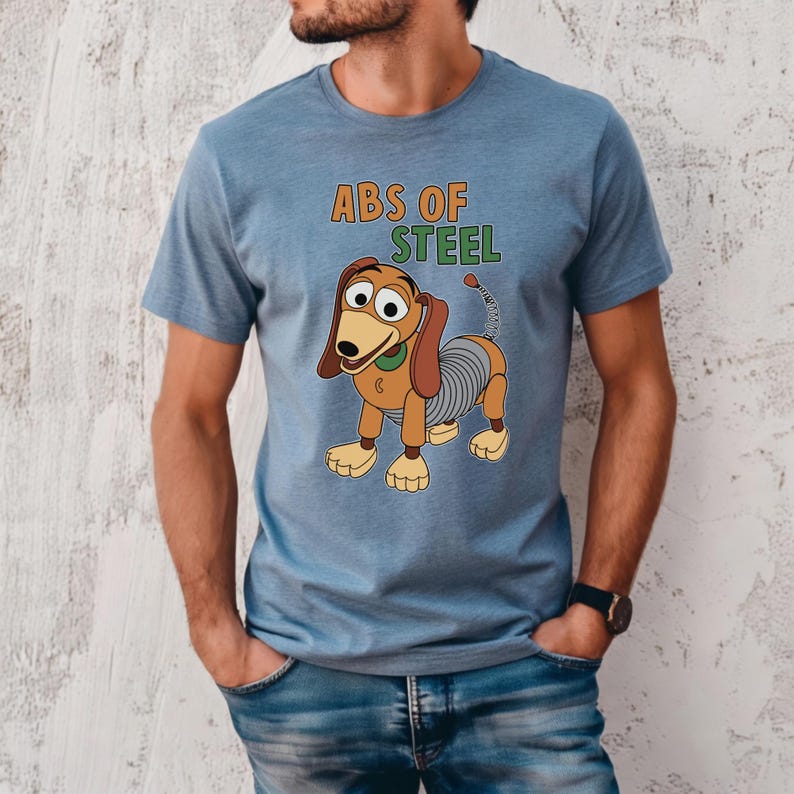 K&ouml;nnte beinhalten: Hellblaues T-Shirt mit einem Cartoon-Bild von Slinky Dog und dem Text "ABS OF STEEL" in Orange und Gr&uuml;n. Der Hund hat braune Ohren, einen gr&uuml;nen Kragen und einen federartigen K&ouml;rper. Das T-Shirt ist aus weichem Material.