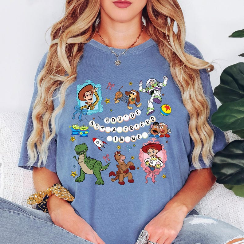 May include: A blue t-shirt with a colourful design featuring cartoon characters and the text "YOU'VE GOT A FRIEND IN ME". The design includes a cowboy, a space ranger, a dinosaur, and other characters.