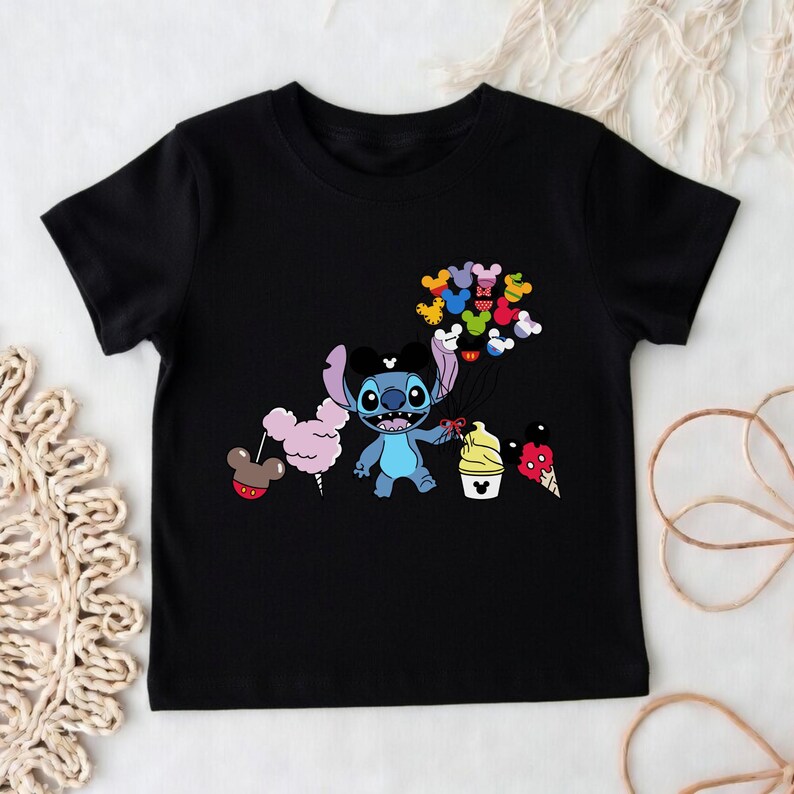 Puede incluir: Camiseta negra con un personaje de dibujos animados sosteniendo globos de Mickey Mouse y golosinas. El personaje azul tiene ojos grandes y un lazo negro. Las golosinas incluyen algod&oacute;n de az&uacute;car, helado y una piruleta con forma de Mickey Mouse.