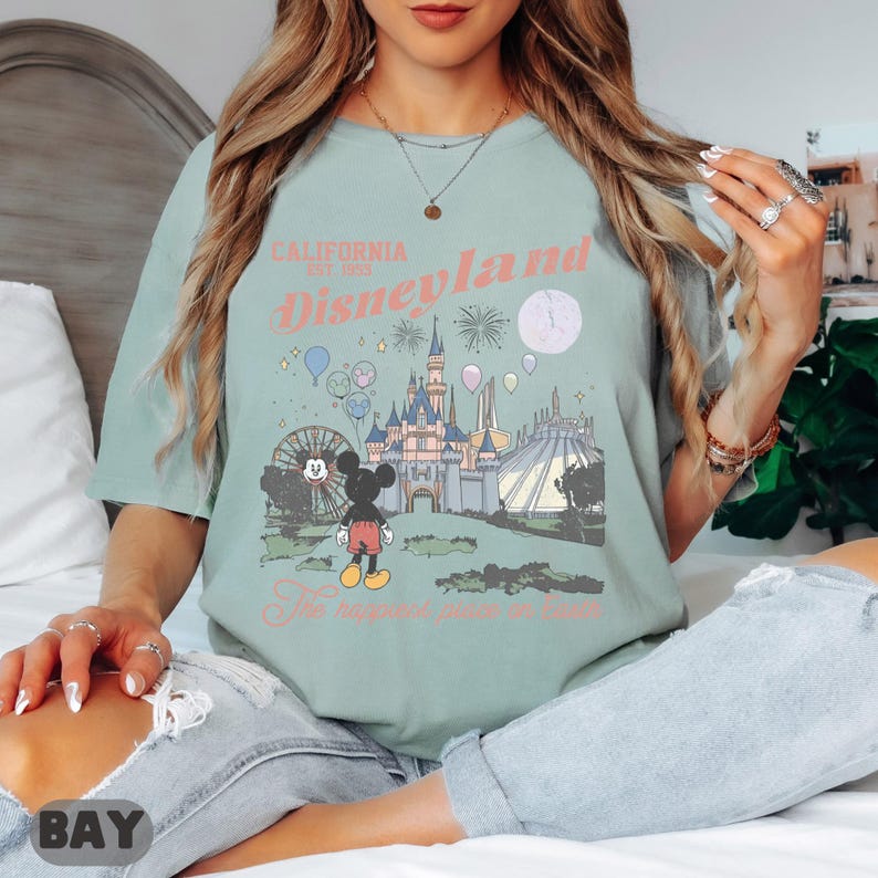 May include: Sage green t-shirt featuring a Disneyland graphic. The design includes a castle, Mickey Mouse, fireworks, and the text "California Disneyland Est. 1955" and "The happiest place on Earth".