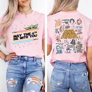 以下が含まれることがあります： ピンク色のTシャツ。ベビーヨーダ、ライトセーバー、そして「MAY THE 4TH BE WITH YOU」の文字が描かれています。背中にはスターウォーズのキャラクターと引用句が描かれています。このTシャツはクルーネックです。
