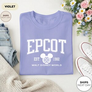 May include: A violet-colored t-shirt with the word "EPCOT" in large white block letters. Below the word "EPCOT" is "EST. 1982" and "WALT DISNEY WORLD". The shirt is folded and next to a pair of white sneakers and a pair of blue jeans.