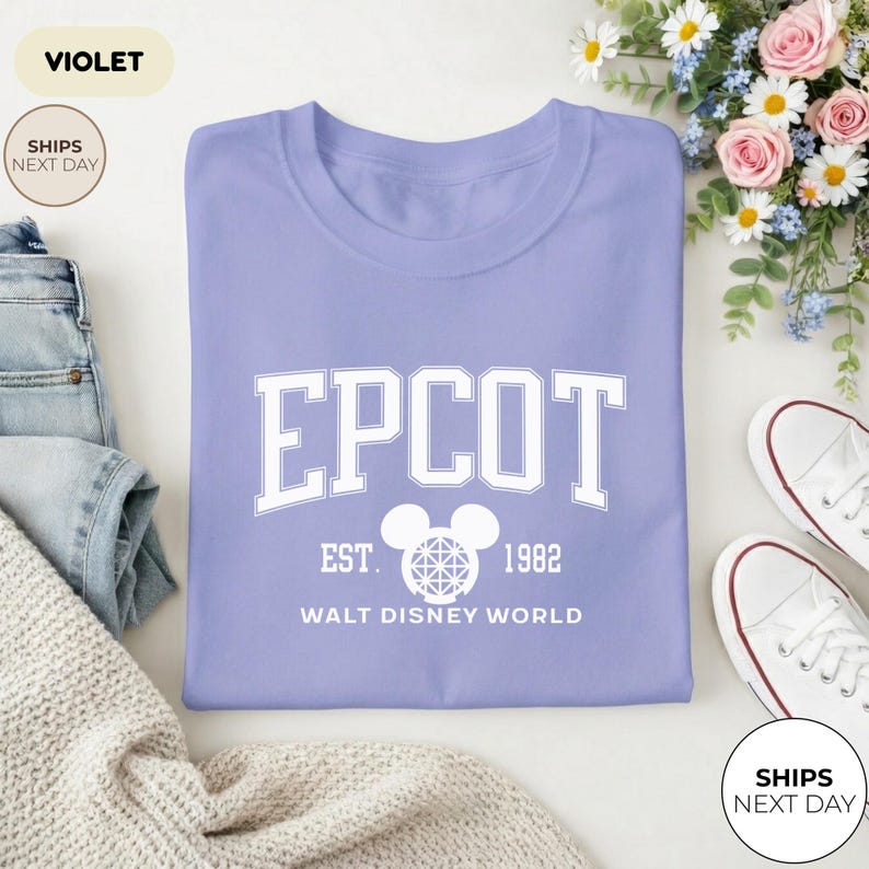 May include: A violet-colored t-shirt with the word "EPCOT" in large white block letters. Below the word "EPCOT" is "EST. 1982" and "WALT DISNEY WORLD". The shirt is folded and next to a pair of white sneakers and a pair of blue jeans.