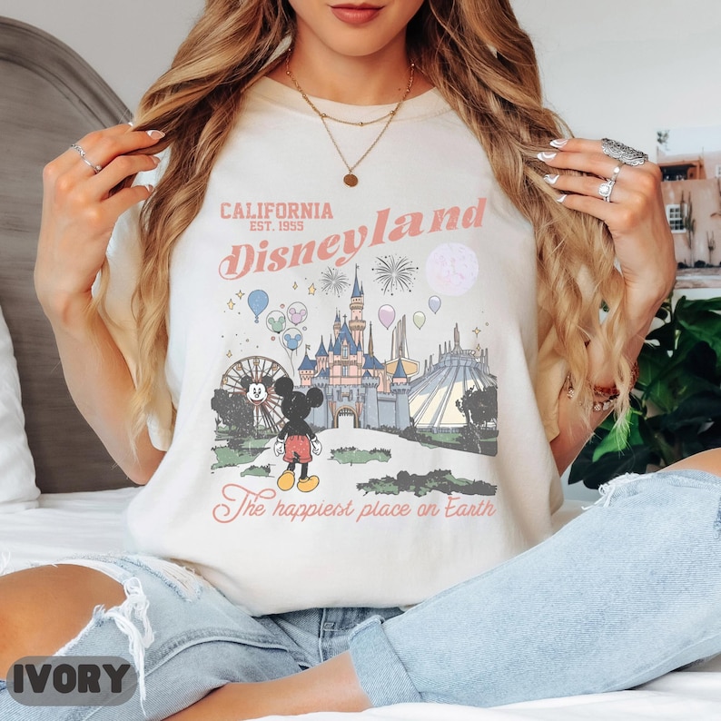 May include: Ivory-colored t-shirt featuring a Disneyland graphic. The design includes the words "California Est. 1955 Disneyland" in pink script, along with an illustration of the castle, Mickey Mouse, and other park elements. The phrase "The happiest place on Earth" is also included.