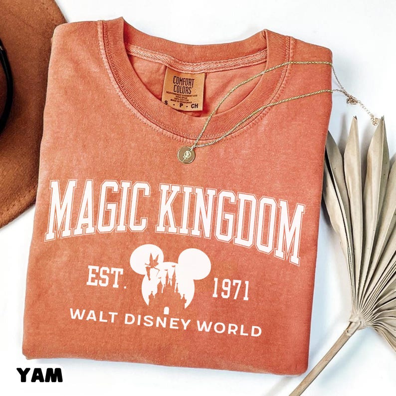 May include: Coral-colored t-shirt with "MAGIC KINGDOM" in white block letters. Below, it reads "EST. 1971 WALT DISNEY WORLD" with a Mickey Mouse silhouette. A gold necklace and a dried palm leaf are also visible.