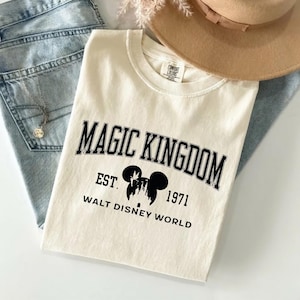 May include: Cream-colored t-shirt with the words "MAGIC KINGDOM" in black, arched text. Below, it reads "EST. 1971 WALT DISNEY WORLD" with a Mickey Mouse silhouette. The shirt is laid on top of blue jeans and a tan hat.