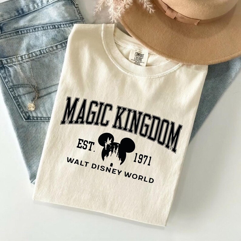 May include: Cream-colored t-shirt with the words "MAGIC KINGDOM" in black, arched text. Below, it reads "EST. 1971 WALT DISNEY WORLD" with a Mickey Mouse silhouette. The shirt is laid on top of blue jeans and a tan hat.