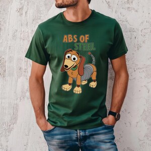 K&ouml;nnte beinhalten: Gr&uuml;nes T-Shirt mit einem Cartoon-Bild von Slinky Dog und dem Text "ABS OF STEEL" in Orange und Gr&uuml;n. Der Hund hat braune Ohren, einen gr&uuml;nen Kragen und einen spiralf&ouml;rmigen K&ouml;rper. Das Shirt ist aus weichem Material.