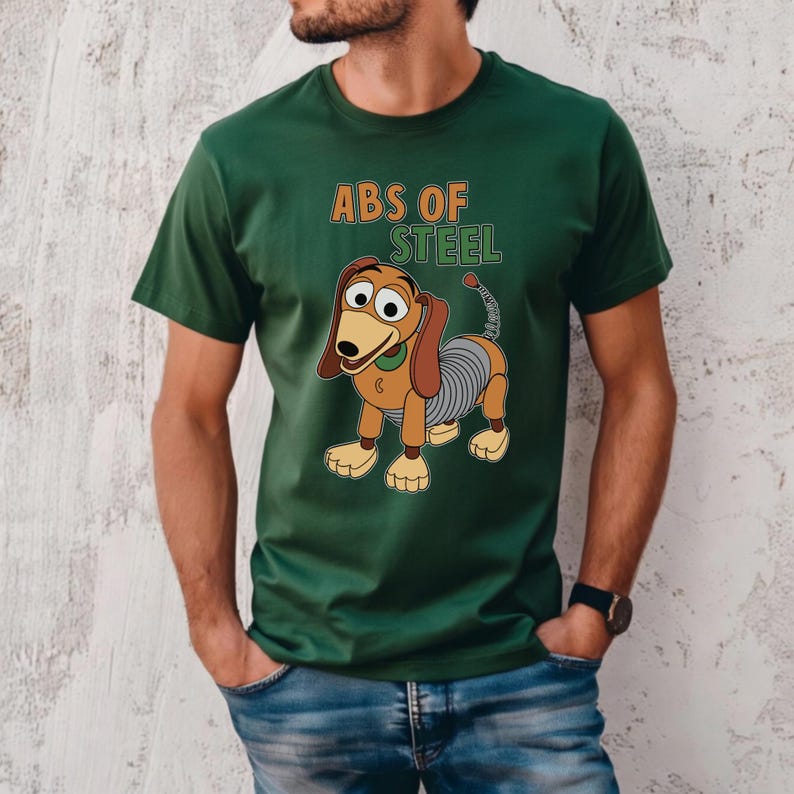 K&ouml;nnte beinhalten: Gr&uuml;nes T-Shirt mit einem Cartoon-Bild von Slinky Dog und dem Text "ABS OF STEEL" in Orange und Gr&uuml;n. Der Hund hat braune Ohren, einen gr&uuml;nen Kragen und einen spiralf&ouml;rmigen K&ouml;rper. Das Shirt ist aus weichem Material.