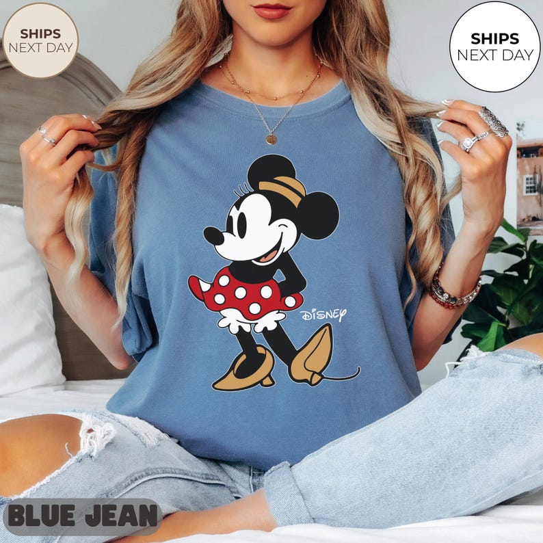 Puede incluir: Camiseta azul claro con un dise&ntilde;o de Minnie Mouse. El personaje de dibujos animados est&aacute; representado con una falda roja de lunares, una parte superior negra y zapatos amarillos. La palabra "Disney" est&aacute; impresa debajo.
