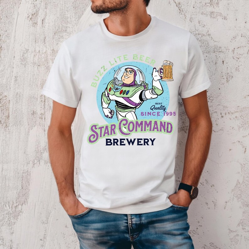 May include: A white t-shirt featuring a graphic of a space ranger character holding a beer mug inside a blue circle. The text reads BUZZ LITE BEER, BEST QUALITY SINCE 1995, STAR COMMAND, and BREWERY in green, blue, and black lettering.
