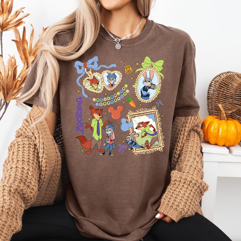 Może przedstawiać: Brązowa koszulka z r&oacute;żnymi obrazkami z kresk&oacute;wek i napisem "Zootopia" w kolorze fioletowym. Koszulka przedstawia postacie takie jak Nick Wilde i Judy Hopps w oprawionych portretach i innych zabawnych wzorach. Zabawna i kolorowa koszulka graficzna.