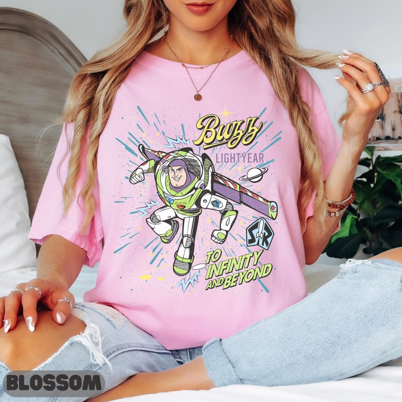 May include: Pink t-shirt featuring a graphic of Buzz Lightyear from Toy Story. The design includes the text "Buzz Lightyear" and "To Infinity and Beyond." The character is depicted in his signature green, white, and purple spacesuit, with a planet and starbursts.