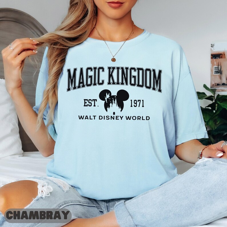 May include: Light blue t-shirt with the text "MAGIC KINGDOM" above "EST. 1971 WALT DISNEY WORLD" and a silhouette of Mickey Mouse ears and a castle. The shirt is a light blue color.