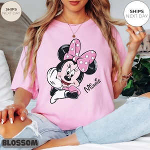 May include: A light pink t-shirt featuring a cartoon image of Minnie Mouse. Minnie Mouse is depicted with a pink polka-dot bow, black ears, and a pink dress. The text "Mr. Minnie" is printed in black on the shirt.