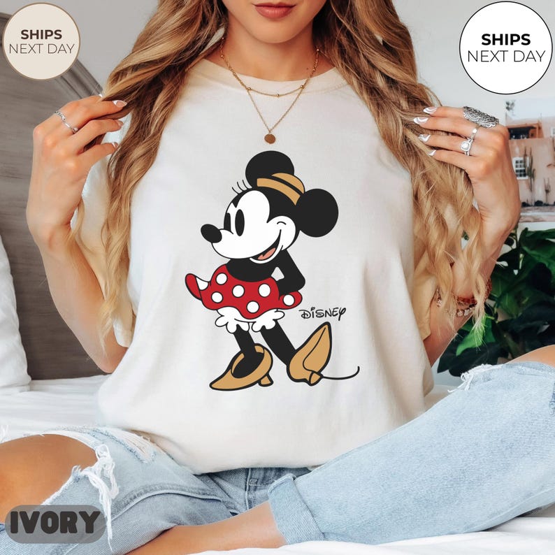 Puede incluir: Camiseta de color marfil con un dibujo de Minnie Mouse. El personaje de dibujos animados lleva una falda roja de lunares y zapatos amarillos. El logotipo de Disney est&aacute; impreso debajo. La camiseta tiene cuello redondo y mangas cortas.
