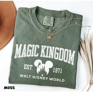 May include: A moss green t-shirt with the words "MAGIC KINGDOM" in white block letters. Below the text is "EST. 1971 WALT DISNEY WORLD" with a Mickey Mouse silhouette. A gold necklace with a pendant rests on the shirt.