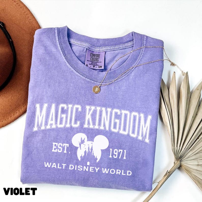 May include: A violet Comfort Colors t-shirt with the words "MAGIC KINGDOM" in white block letters. Below the text is "EST. 1971" and "WALT DISNEY WORLD" in white. A gold necklace with a pendant rests on the shirt.