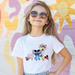 Puede incluir: Una camiseta blanca con un personaje de dibujos animados sosteniendo globos y golosinas. El personaje es azul con orejas grandes y un sombrero negro. La camiseta la lleva una persona con pelo largo y rubio y gafas de sol redondas.
