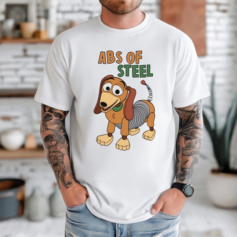 K&ouml;nnte beinhalten: Wei&szlig;es T-Shirt mit einer Cartoon-Illustration eines Dackelhundes und dem Text "ABS OF STEEL". Der Hund hat einen federartigen K&ouml;rper. Der Text ist in Orange und Gr&uuml;n. Ein l&auml;ssiges Kleidungsst&uuml;ck.