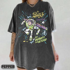 Może przedstawiać: Ciemnoszara koszulka z grafiką Buzza Astrala w pozycji lotu, z napisami "Buzz Lightyear" i "To Infinity and Beyond". Projekt zawiera tło o tematyce kosmicznej z rozbłyskami gwiazd i liniami.