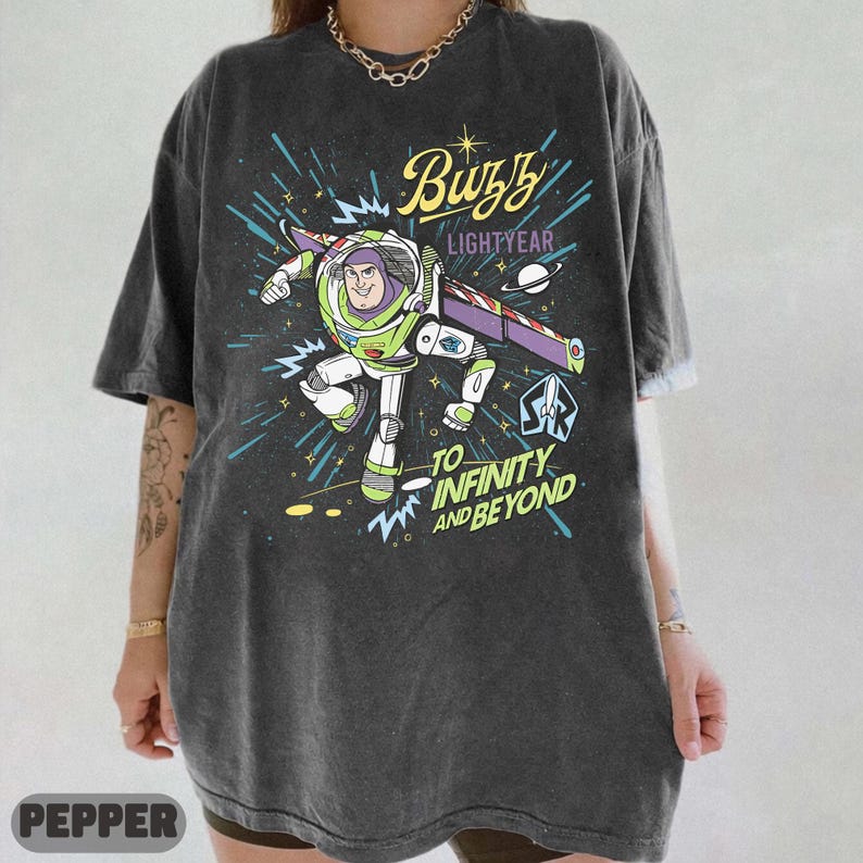 Może przedstawiać: Ciemnoszara koszulka z grafiką Buzza Astrala w pozycji lotu, z napisami "Buzz Lightyear" i "To Infinity and Beyond". Projekt zawiera tło o tematyce kosmicznej z rozbłyskami gwiazd i liniami.