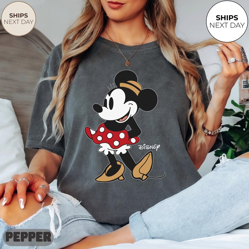 Puede incluir: Camiseta gris oscuro con un dise&ntilde;o de Minnie Mouse. El personaje de dibujos animados est&aacute; representado en negro, blanco, rojo y amarillo, con la palabra "Disney" impresa debajo. La camiseta tiene un aspecto vintage y desgastado.