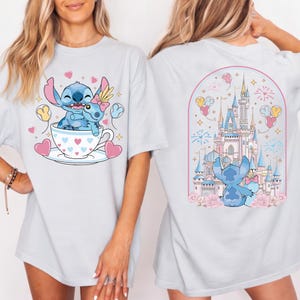 May include: Light blue t-shirt featuring a cartoon Stitch design. The front shows Stitch in a teacup with hearts and balloons. The back displays Stitch in front of a castle with fireworks and flowers. A fun, whimsical design.