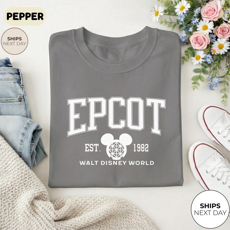 May include: A gray crew neck t-shirt with the word "EPCOT" in white block letters. Below the word "EPCOT" is the text "EST. 1982 WALT DISNEY WORLD" in white. A Mickey Mouse head graphic is between "EST." and "1982".