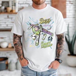 May include: White t-shirt featuring a colorful graphic of Buzz Lightyear from Toy Story. The design includes the text "Buzz Lightyear" and "To Infinity and Beyond" with a space-themed background. The shirt has short sleeves.