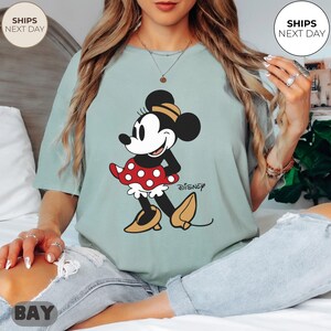 Puede incluir: Camiseta verde claro con un gr&aacute;fico de Minnie Mouse. El personaje de dibujos animados est&aacute; representado con una falda roja de lunares, una parte superior negra y zapatos amarillos. El logotipo de Disney est&aacute; impreso debajo.