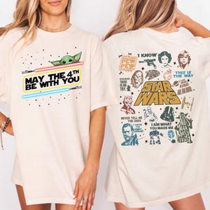 以下が含まれることがあります： スターウォーズのグラフィックが特徴のクリーム色のTシャツ。「May the 4th Be With You」の文字と、ヨーダとライトセーバーが前面にプリントされています。背面には、スターウォーズシリーズの様々なキャラクターと引用句が描かれています。
