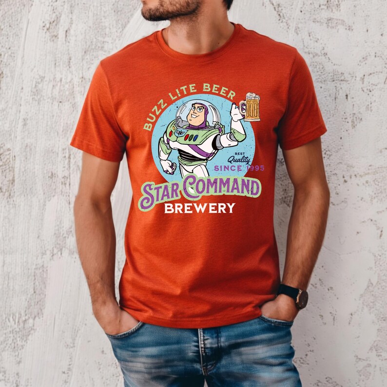 May include: A burnt orange short-sleeve t-shirt featuring a graphic of a space ranger character holding a beer mug. The text reads BUZZ LITE BEER, BEST QUALITY SINCE 1995, STAR COMMAND BREWERY. The design is set against a light blue circular background.