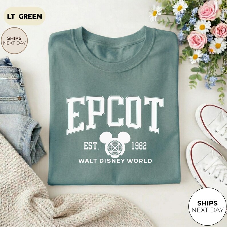 May include: A light green t-shirt with the word "EPCOT" in white block letters. Below the word "EPCOT" is a Mickey Mouse head with a geometric design, the text "EST. 1982" and "WALT DISNEY WORLD". The shirt is folded with a pair of white sneakers.